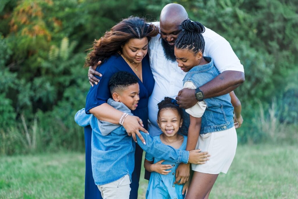 A happy family hugging each other in a circle outdoors, 3 kids and two adults.