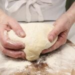 A chef in the kitchen kneading a large piece of dough with flour on his hands.
