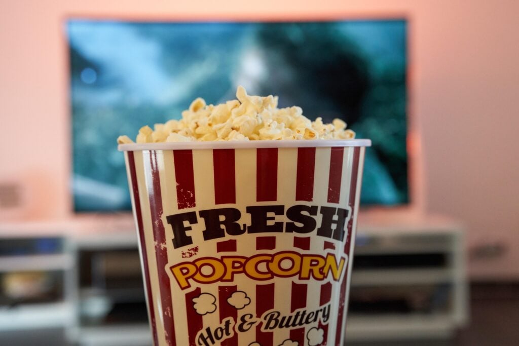 A TV is blurry in the background, with a container of popcorn front and center to signify home entertainment.