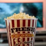 A TV is blurry in the background, with a container of popcorn front and center to signify home entertainment.