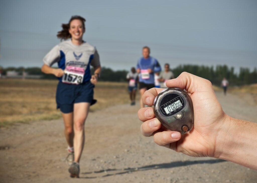 A marathon with running woman in the lead, hand with stopwatch in the foreground).