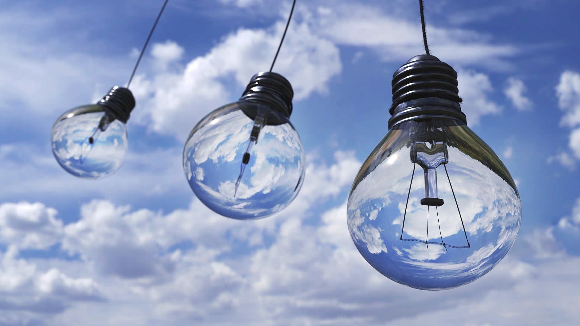 Three light bulbs on cords swinging in the blue sky, reflections of the sky and clouds can be seen inside the bulbs.